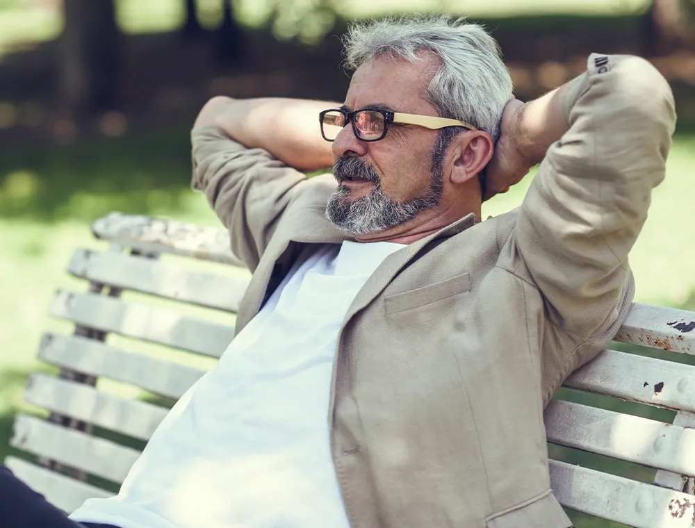 A mature man with glasses sitting comfortably on a bench outdoors with hands behind his head, enjoying pain-free relaxation.