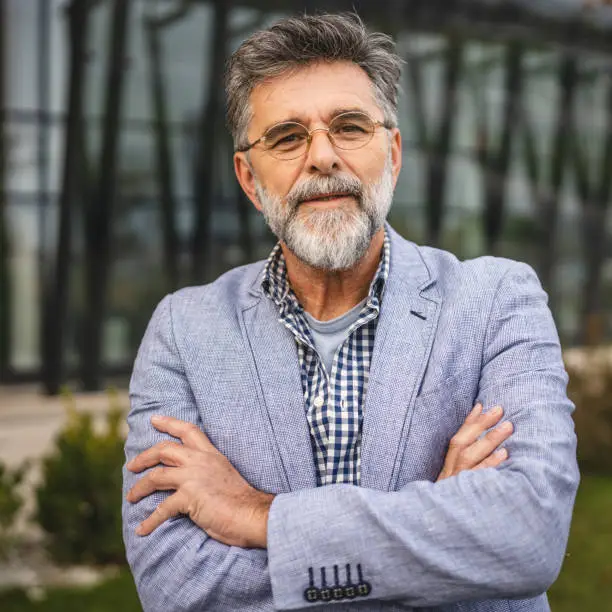Portrait of a smiling senior man with a beard and grey sweater crossing his arms, showcasing physical health and daily wellness.